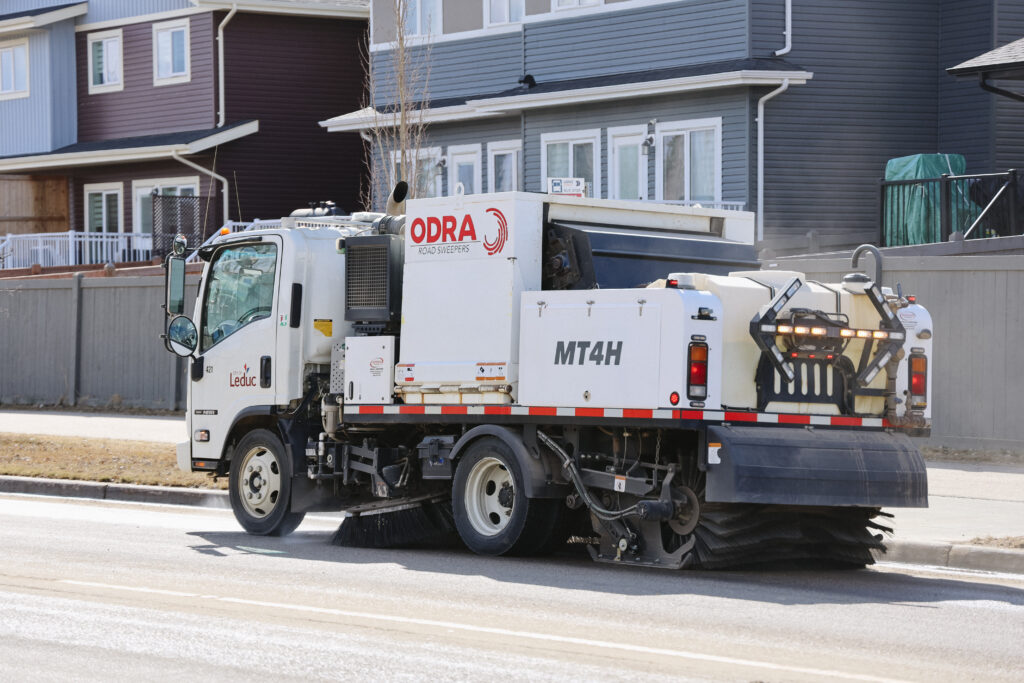 City of Leduc street sweeper truck sweeping debris from a residential road during daylight hours, with modern homes and backyard fences lining the street.