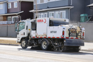 City of Leduc street sweeper truck sweeping debris from a residential road during daylight hours, with modern homes and backyard fences lining the street.