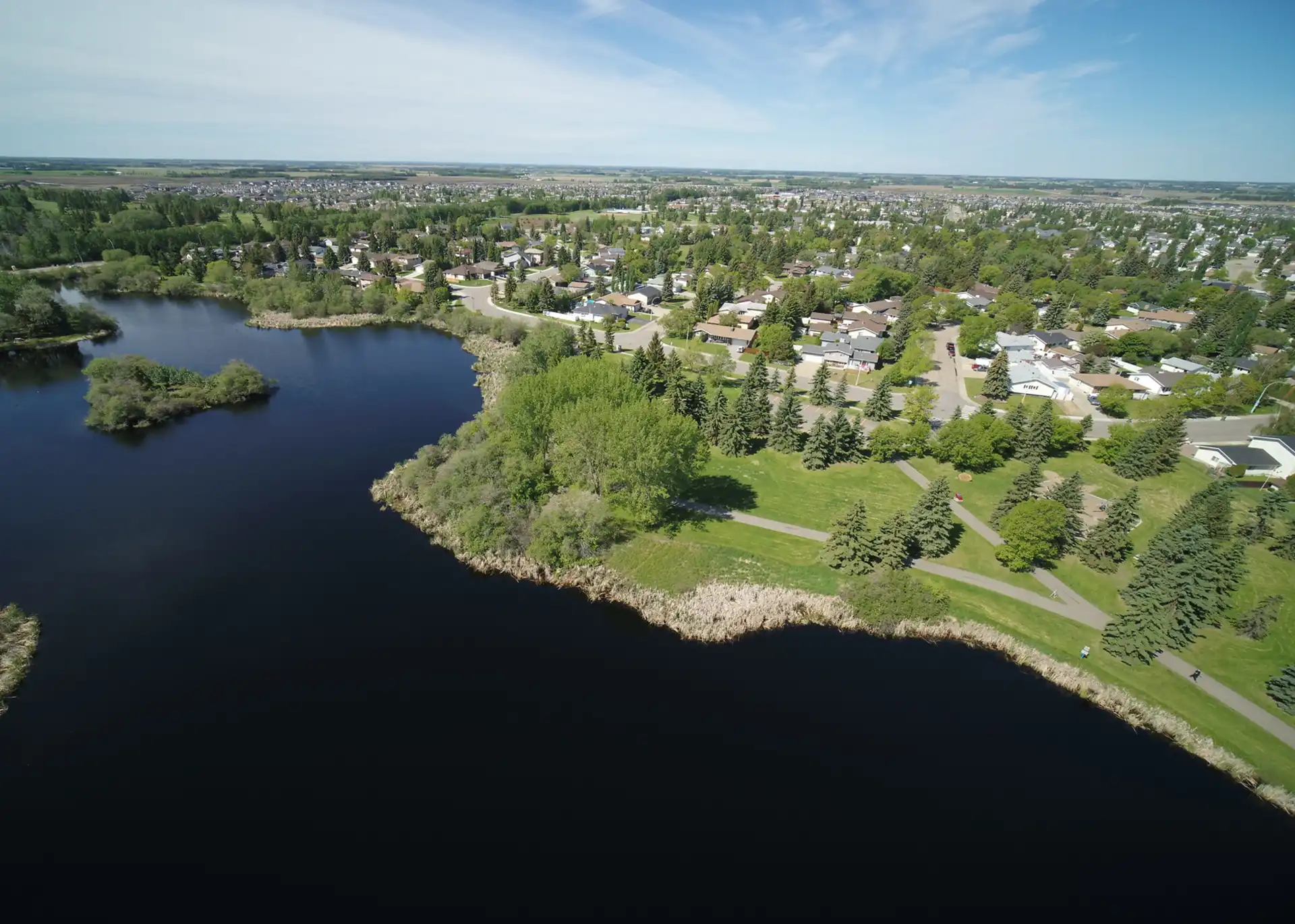 Aerial view of a lakeside community with lush greenery, residential houses, and a winding path along the shoreline under a partly cloudy sky.