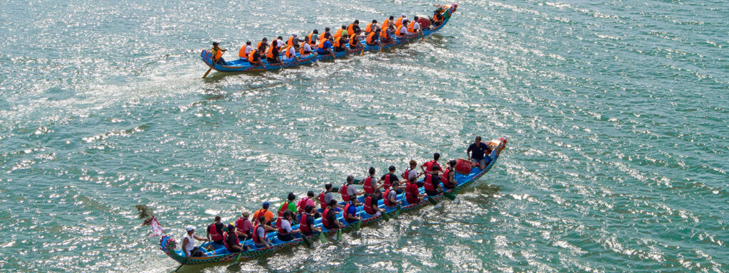Aerial view of two dragon boats on sparkling blue-green water, with teams of paddlers wearing brightly coloured life jackets rowing in unison.