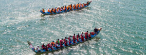 Aerial view of two dragon boats on sparkling blue-green water, with teams of paddlers wearing brightly coloured life jackets rowing in unison.