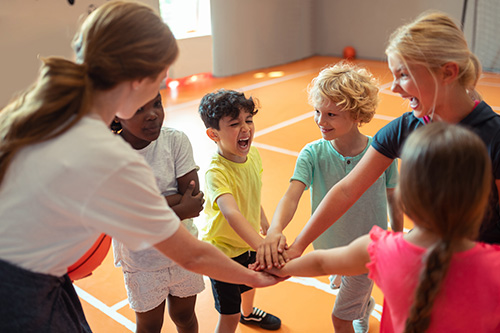 Children and a program leader participate in a group activity, placing their hands together in a gymnasium during a Kids' Night Out program.